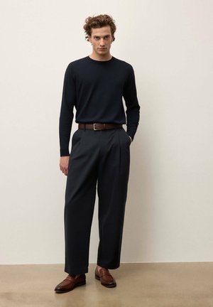 A man stands wearing a black long-sleeve shirt, loose black trousers, brown leather belt, and brown leather loafers against a plain background.