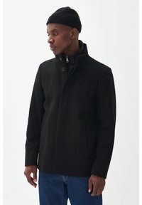 Cappotto nero in lana con colletto alto, due tasche frontali e chiusura con zip, abbinato a un berretto nero a costine. Finitura in tessuto strutturato.
