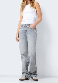 Personne portant un jean droit gris clair, un débardeur blanc et des baskets, debout avec une main dans la poche.