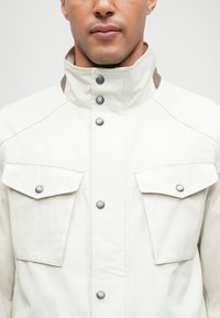 Chaqueta beige clara con un alto collar, botones a presión y dos bolsillos frontales. Presenta detalles en marrón contrastante en el collar. Textura suave.