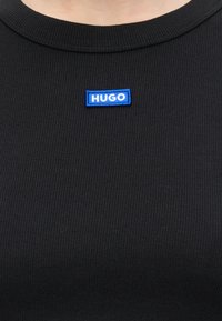 Gros plan sur un tissu à côtes noir avec un petit patch rectangulaire bleu affichant le mot HUGO en lettres blanches.