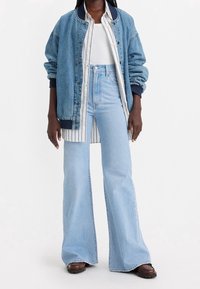 Person med ljusblå vida jeans, vit ribbad topp, randig knappad skjorta, överstora denimjacka och bruna skor mot vit bakgrund.