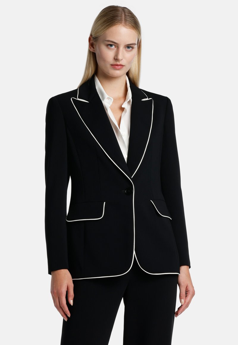Blazer nero con piping bianco, chiusura a un bottone, due tasche frontali, vestibilità sartoriale e texture del tessuto liscia. Abbinato a una camicia bianca.