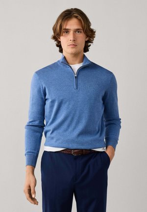 Joven con un suéter azul claro con cremallera sobre una camisa blanca, pantalones azul marino y un cinturón marrón tejido, de pie frente a un fondo liso.