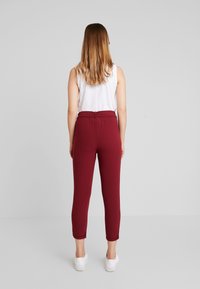 Bordeauxrode, rekbare broek met een aangesloten taille en zakken tot de enkels, met een elastische tailleband en zijnaad. Gecombineerd met witte schoenen.
