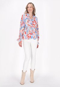 Bluză colorată cu un model floral în roșu, roz și albastru, având un decolteu cu nasturi și mâneci volan, asortată cu pantaloni albi și cizme deschise.