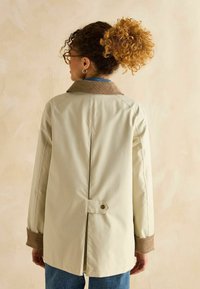 Cămașă bej cu guler din velur maro, fanta la spate și detalii cu nasturi. Prezintă mâneci lungi și o croială lejeră. Modelul poartă blugi albaștri.