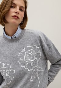 Grijze trui met witte bloemige borduursels, ronde hals en textuur gebreide stof. Gelaagd over een blauw-wit gestreepte shirt.