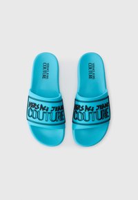 Blue rubber slide sandals feature a textured upper and a black printed logo "VERSACE JEANS COUTURE" on the straps. Flat sole.