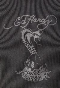 Eine beschlungene Schlange erhebt sich aus einem gesprungenen Schädel mit sichtbaren Reißzähnen, unter dem geschwungenen "Ed Hardy"-Schriftzug auf einem dunkel strukturierten Stoff.