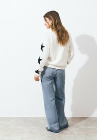White knit sweater with black star patterns on sleeves, paired with blue wide-leg jeans. Sweater has a ribbed hem and cuffs.