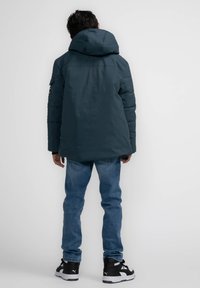 Mörkblå vadderad jacka med stor huva, texturerat material, sidfickor och en logotypapplikation på vänster ärm. Tillsammans med blå jeans och svarta sneakers.