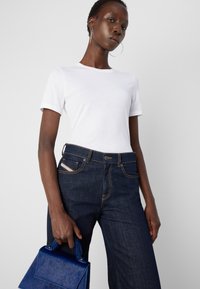 Femme portant une chemise blanche à manches courtes et un jean foncé, tenant un petit sac à main bleu texturé sur un fond clair uni.