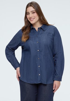 Pitkät ruskeat hiukset omaava nainen, joka pukeutunut napitettuun tummansiniseen farkkupaitaan ja vastaaviin tummansinisiin farkkuihin, hymyilee ja pitää kättään lanteillaan.