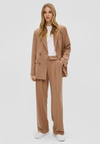 Costume beige à la coupe ajustée composé d'un blazer croisé et de pantalons à jambe large, associé à un t-shirt blanc et des baskets blanches.