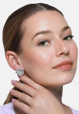 Donna con pelle chiara e occhi verdi che indossa grandi orecchini d’argento a forma di conchiglia, sorride dolcemente, con la mano che sfiora delicatamente il collo.