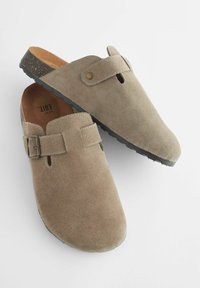 Sabots en daim beige avec un bout arrondi, un détail de boucle et une semelle en caoutchouc noir texturé. Dispose d'un intérieur doux et doublé.