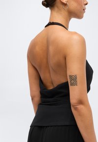 Γυναίκα με μαύρο τοπ χωρίς πλάτη που δείχνει την ακάλυπτη πλάτη της και ένα τετράγωνο τατουάζ με λουλούδια στο πάνω αριστερό της μπράτσο.