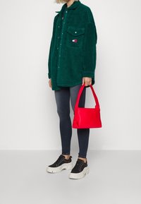 Person som bär en grön fleecejacka, mörkgrå skinny jeans, svarta och vita platåskor, och håller en liten ljusröd handväska.