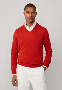 Muž v červeném svetr s hlubokým výstřihem přes bílou košili s kostkovaným vzorem, v kombinaci s bílými kalhotami, stojící na jednobarevném pozadí.