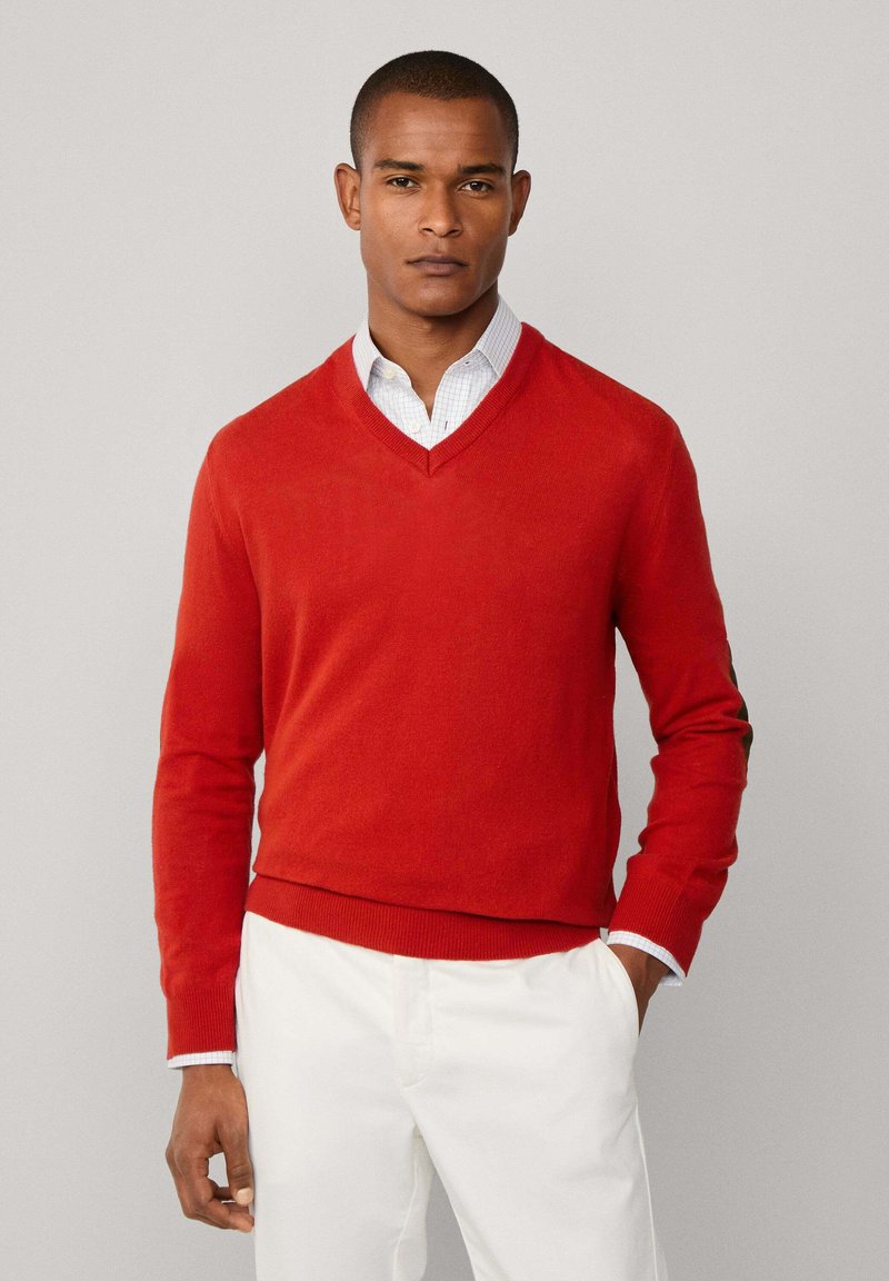 Muž v červeném svetr s hlubokým výstřihem přes bílou košili s kostkovaným vzorem, v kombinaci s bílými kalhotami, stojící na jednobarevném pozadí.