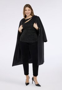 Schwarzer langer Mantel über einem gepunkteten Oberteil, kombiniert mit schwarzen Samthosen und schwarzen Absatzschuhen. Glatte Texturen und ein figurbetontes Design mit Ausschnittdetail.