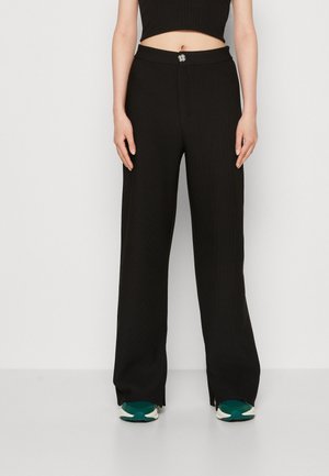 Vrouw draagt zwarte, hoog getailleerde wijde broek met knopen aan de voorkant, gecombineerd met turquoise en witte sneakers en een zwart cropped top.