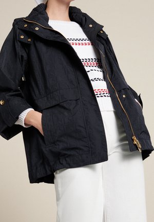 Femme portant une veste noire zippée avec des boutons dorés et un pull blanc avec des rayures rouges et noires, associée à un pantalon blanc.