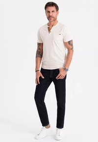 Hvid T-shirt med korte ærmer og henley-design, med brystlomme og brune knapdetaljer, parret med mørke jeans og hvide sneakers.