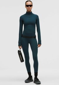 Kobieta w turkusowym stroju sportowym, czarnych sneakersach i okularach przeciwsłonecznych trzyma czarną butelkę na wodę z napisem "lululemon".