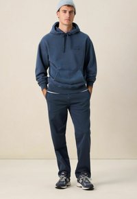 Marineblauwe hoodie met een voorzak, gecombineerd met marineblauwe broek en grijze sneakers. De outfit heeft een casual pasvorm en een zachte textuur.