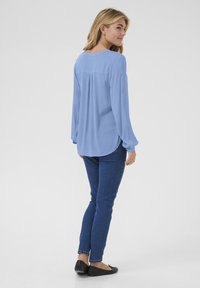 Blouse bleu clair à manches longues avec un col rond, coupe décontractée et fentes sur les côtés, associée à un jean bleu foncé et des ballerines noires.