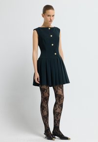 Robe sans manches vert foncé avec des boutons dorés, jupe plissée. Des collants noirs à motif dentelle et des ballerines noires pointues complètent la tenue.