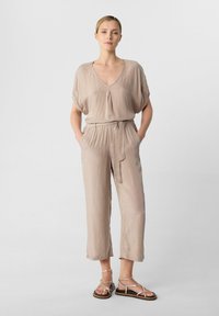 Beige, kurzärmliger Overall mit V-Ausschnitt, elastischem Bund und Seitentaschen, ergänzt durch goldene Sandalen mit Riemchen. Glattes, leichtes Material.