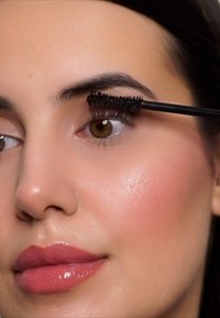 Maschera nera applicata con un pennello su lunghe ciglia. Il viso ha una pelle liscia, labbra lucide e un leggero fard sulle guance.