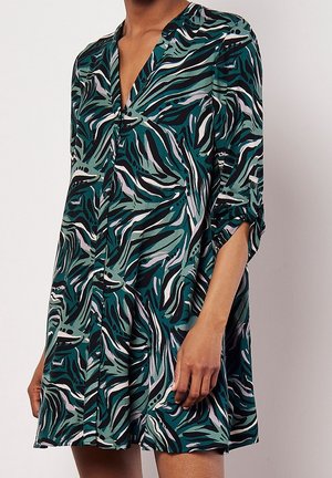 Kvinde iført en løs, langærmet kjole med et abstrakt bladmønster i grøn, sort og hvid samt ærmer, der er rullet op.