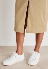 Beige Baumwollrock mit einem Schlitz vorne, kombiniert mit weißen Leinen-Sneakern mit runder Spitze und Schnürdesign auf einem neutralen Boden.