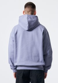 Lila hoodie visad bakifrån, med en stor huva, lös passform och textilstruktur. Kombinerad med svarta byxor.