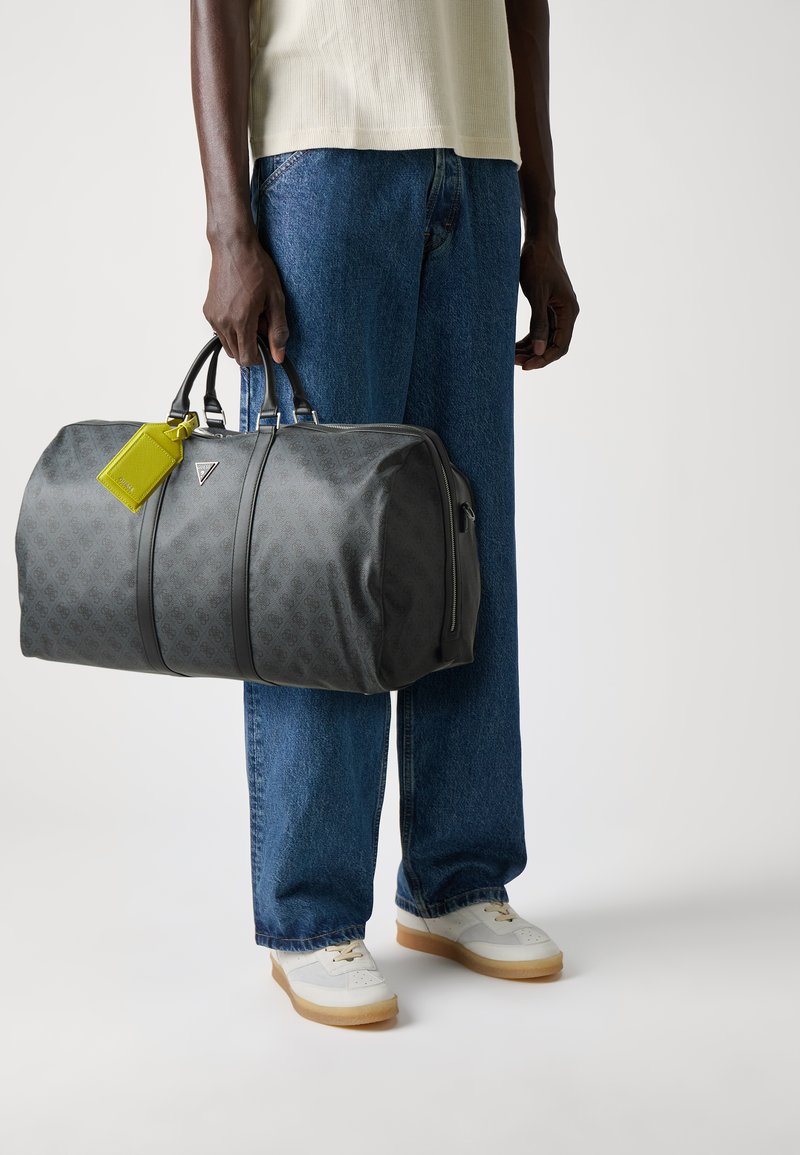 Sac de sport gris avec un motif monogramme discret, accents en cuir noir, quincaillerie en argent et étiquette de bagages jaune vif.