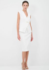 Blazer senza maniche bianco con scollatura a V profonda e un accento dorato, abbinato a una gonna a matita bianca fino al ginocchio e tacchi beige a cinturino alla caviglia.