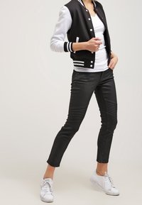 Svart varsityjacka med vita ärmar, randiga muddar; vit V-ringad t-shirt; svarta slim-fit-byxor med ribbad textur; vita sneakers.