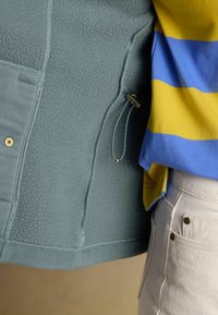 Veste en polaire bleu gris avec un cordon de serrage à côté d'une personne portant une chemise rayée jaune et bleue ainsi qu'un pantalon beige clair.