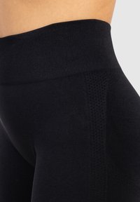 Zwarte sportleggingen met een brede tailleband en gestructureerde zijpanelen, benadrukt tegen een neutrale achtergrond.