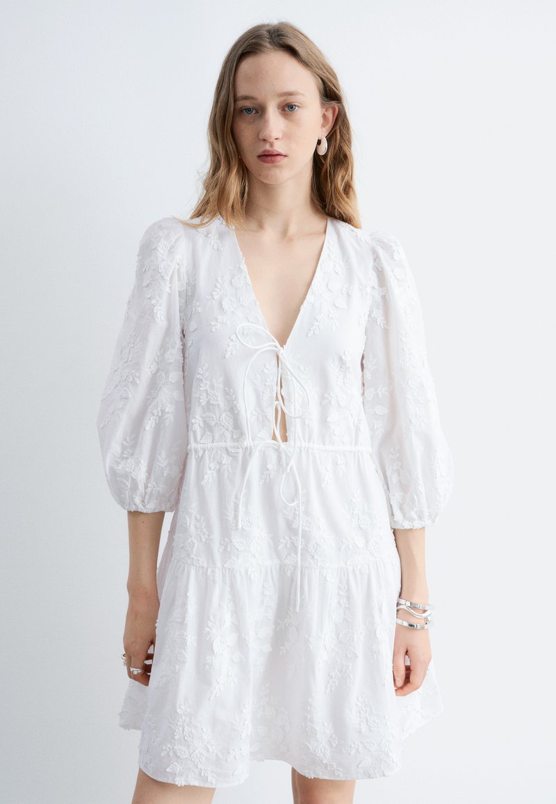 Femme portant une robe blanche brodée de fleurs avec des manches trois-quarts bouffantes et un profond décolleté en V noué à l'avant, accessoirisée de bracelets argentés.