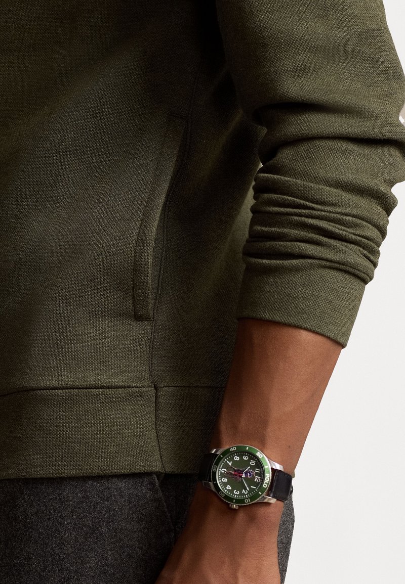 Olivegrüner strukturierter Sweatshirt mit einer Fronttasche, kombiniert mit einer runden Uhr mit grünem Rahmen, schwarzem Armband und kontrastierenden Ziffernblatt-Details.