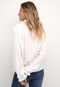 White blouse with gathered sleeves, smooth texture, and subtle sheen, paired with blue jeans. The shirt features a back yoke detail.