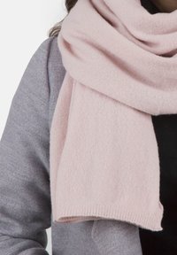 Une écharpe en cachemire rose pâle drape doucement, présentant une texture fine et des bords lisses, associée à un pull gris en arrière-plan.