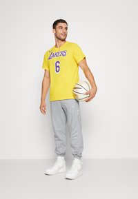 Cămașă galbenă cu mâneci scurte, având textul "LAKERS" și numărul 6, asortată cu pantaloni de trening gri și adidași albi cu glezna înaltă, ținând o minge de baschet.