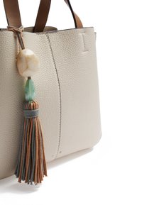 Sac cabas beige texturé avec poignées en cuir marron. Présente un pompon décoratif avec des accents verts et beiges, ajoutant un contraste de couleurs.