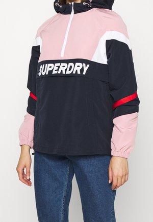 Personne portant une veste zippée rose, noire et blanche avec "SUPERDRY" sur la poitrine et un jean bleu.